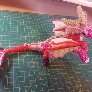 May include: A crocheted pink, red and beige dragon with wings and a long tail. The dragon is sitting on a green cutting mat.