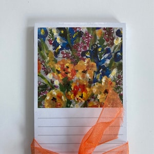 May include: A notepad with a floral painting on top, featuring orange, yellow, blue, and purple flowers. The notepad is tied with an orange ribbon. The lined paper is ready for notes.