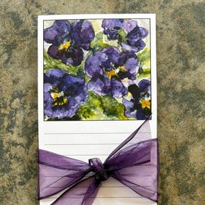 May include: A notepad with a purple ribbon tied around it. The notepad has a watercolor illustration of purple pansies on the top. The notepad has lined paper.