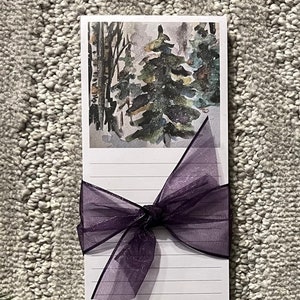 May include: A white notepad with lined paper and a purple ribbon tied around it. The notepad has a watercolor illustration of a snowy forest scene on the front.