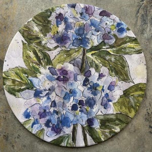 May include: Round decorative plate featuring a watercolor painting of blue and purple hydrangeas with green leaves. The floral design is set against a white background, creating a soft, artistic aesthetic. The plate is ideal for home decor.