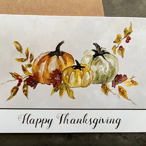 May include: A watercolor painting of three pumpkins with fall leaves and berries. The pumpkins are orange, yellow, and green. The text "Happy Thanksgiving" is written below the pumpkins.