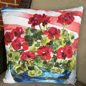May include: Square decorative pillow featuring a watercolor painting of red geraniums in a blue pot. The background has red and white stripes with blue stripes at the top. The pillow is on a neutral-colored surface.