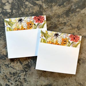 May include: Two white blank note cards with a floral design border featuring yellow, orange, and red flowers with green leaves. The cards are square and sit on a textured, dark gray surface.