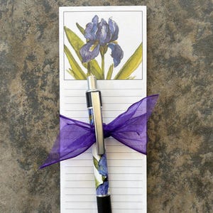 May include: A notepad with a watercolor painting of a purple iris flower and green leaves. A pen with a floral design is placed on the notepad and tied with a purple ribbon. The notepad has lined pages.