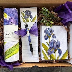 May include: A gift box containing a rolled dish towel, notepad, pen, and a small framed print. The items feature watercolor iris flower designs in shades of purple, blue, and yellow. The dish towel is white with green and yellow stripes. The box is filled with brown packing material and purple tissue paper.