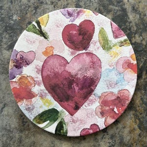 May include: Round coaster with a watercolor design featuring two large burgundy hearts and smaller hearts, surrounded by colorful flowers and green leaves on a white background. The coaster has a floral and heart theme.