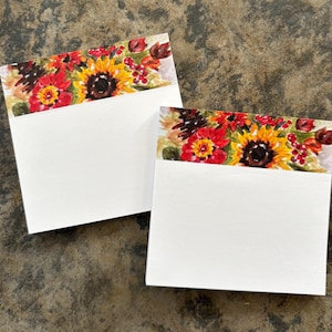 May include: Two square paper notepads featuring a watercolor illustration at the top. The design displays a vibrant arrangement of yellow sunflowers, red flowers, dark brown pinecones, and small red berries against a soft, neutral background.