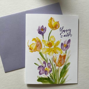 May include: A white ceramic mug with a black handle and the text "But First, Coffee" in a bold, black font. The mug is set against a blurred background, suggesting a cozy setting.