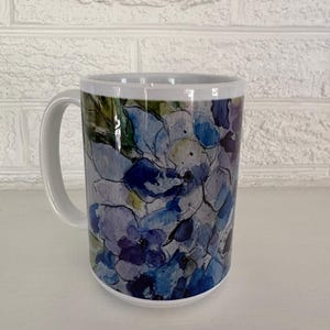 May include: White ceramic mug featuring a watercolor floral design in shades of blue, purple, and green. The mug has a white handle and a glossy finish, suitable for hot or cold beverages.