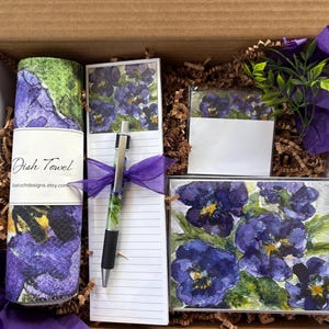 May include: A gift box containing a dish towel, notepad, pen, and notecards. The dish towel is rolled up and wrapped with a white label that reads "Dish Towel". The notepad, pen, and notecards feature a watercolor floral design with purple pansies.
