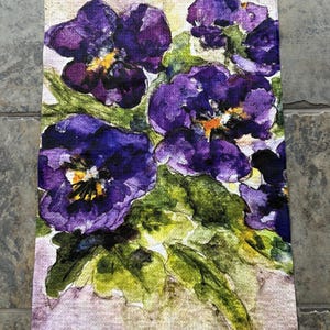 May include: A rectangular kitchen towel featuring a watercolor painting of purple pansies with yellow centers and green leaves. The floral design is set against a light beige background.