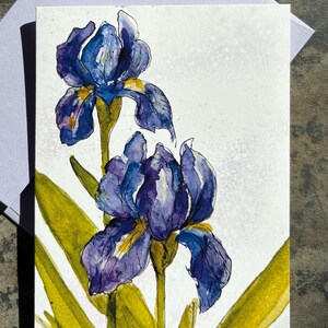 May include: Watercolor painting of two blue and purple iris flowers with yellow-green leaves. The flowers have detailed petals and are set against a white background. The artwork is on a white card, suggesting a greeting card or art print.