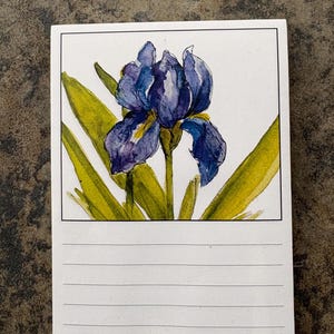 May include: A notepad featuring a watercolor painting of a blue iris flower with yellow and green leaves. The flower is centered on a white background, with lined paper below for notes. The notepad is on a textured, brown surface.