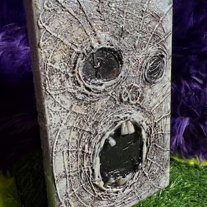May include: A rectangular canvas art piece featuring a textured, distressed grey surface with a sculpted, screaming face design. The face includes two dark, recessed eye sockets and an open mouth revealing several individual white teeth.