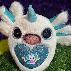 May include: A white plush toy featuring a teal horn, large black glittery eyes, and external gills. The toy has a mouth with visible human-like teeth and a blue glittery heart patch on the front displaying a small cartoon axolotl character.
