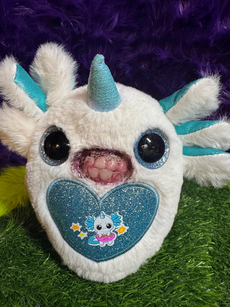 May include: A white plush toy featuring a teal horn, large black glittery eyes, and external gills. The toy has a mouth with visible human-like teeth and a blue glittery heart patch on the front displaying a small cartoon axolotl character.