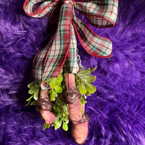 May include: A Halloween-themed decoration featuring three finger-shaped ornaments with a dark brown topping, attached to a green leafy base. A red, green, and white plaid ribbon bow tops the decoration, suspended by a silver chain.