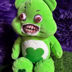 May include: A green plush bear with a white belly featuring a green shamrock. The bear has red, bloodshot eyes and a gruesome, open mouth with visible teeth. The bear's paws also have green heart-shaped details.