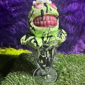 May include: A novelty sundae sculpture with a green, grinning face, pink eyes, and a mouth full of teeth. Black drips cascade down the green face and into the clear glass sundae dish. The sundae is topped with white whipped cream.