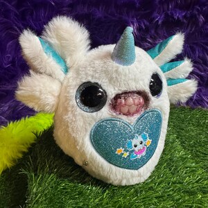 May include: A white plush toy featuring a glittery blue horn, large black eyes, and a mouth with visible teeth. The toy has teal accents on its head appendages and a large, glittery blue heart patch on its chest with a small cartoon graphic.