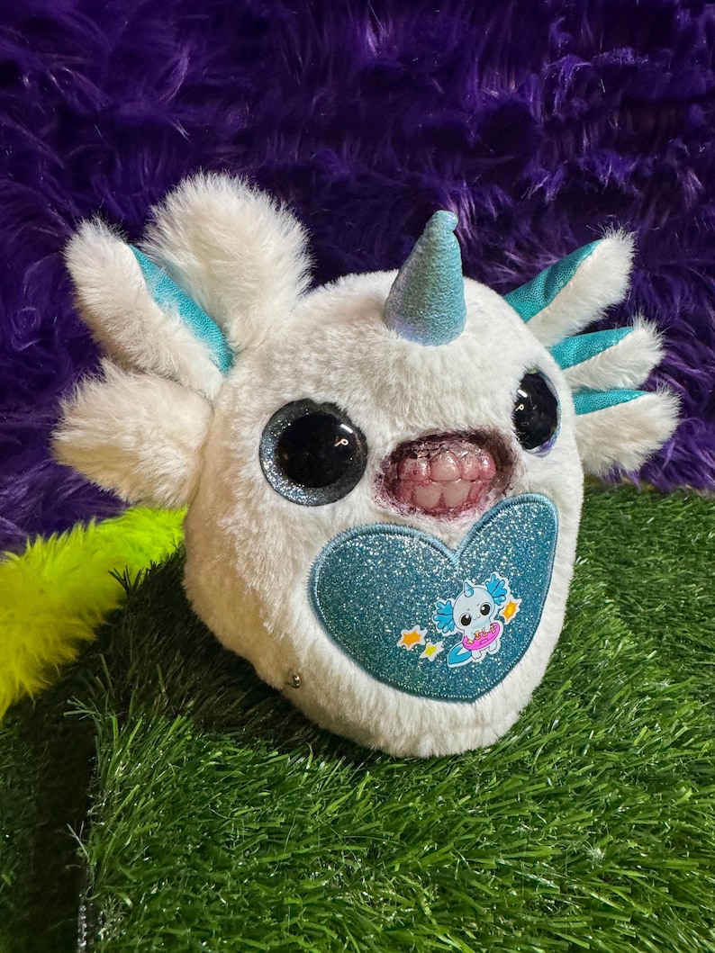 May include: A white plush toy featuring a glittery blue horn, large black eyes, and a mouth with visible teeth. The toy has teal accents on its head appendages and a large, glittery blue heart patch on its chest with a small cartoon graphic.