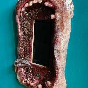 Puede incluir: Una toma de corriente para cargador de teléfono con forma de boca monstruosa con filas de dientes y una apariencia carnosa texturizada.