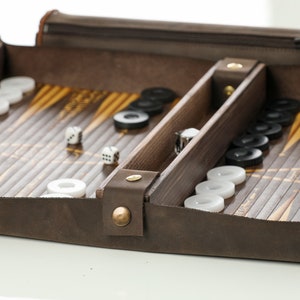 May include: A brown leather travel case with a wooden backgammon board and dice inside. The board has a black and white checkerboard pattern and the case has a zipper closure.