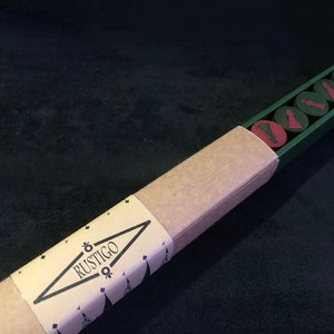 Puede incluir: Una caja de cartón marrón con una tapa verde y la palabra "RUSTIGO" impresa en el lateral. La caja contiene un juego de seis piezas de juego rojas y verdes.