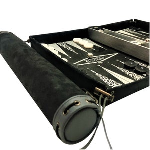 May include: A black backgammon set with a white and black game board, white playing pieces, and black playing pieces. The set comes with a black carrying case with a gray leather strap.