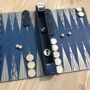 Könnte beinhalten: Ein Backgammon-Spiel mit einem blauen und silbernen Spielbrett, weißen Spielsteinen und Würfeln. Das Brett ist zusammengeklappt und die Würfel zeigen eine 2 und eine 3. Der Punktestand ist 28.