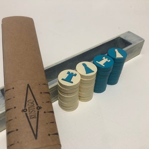 Puede incluir: Una caja de madera con un acabado pintado en azul y blanco contiene pilas de piezas de juego azules y blancas. Las piezas son redondas y tienen un diseño de pieza de ajedrez blanco. La caja tiene una etiqueta de papel marrón con la palabra "RUSTIGO" impresa en ella.