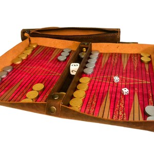 May include: A brown leather backgammon set with a red and white playing surface. The set includes gold and silver game pieces, dice, and a wooden divider. The backgammon board is open, ready for play, and has a zippered pouch.