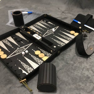May include: A black backgammon set with gold-colored chips and a white and black game board. The set is open and ready to play. The set is in a black leather carrying case with a strap.