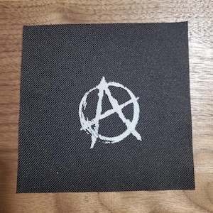May include: A black fabric patch with a white anarchy symbol. The symbol is a circle with an A inside, with lines radiating out from the center.