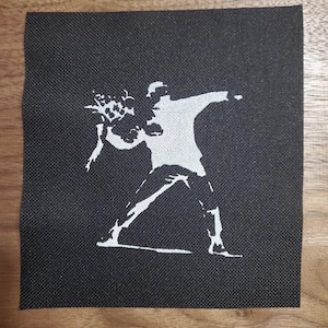 May include: A white silhouette of a person throwing a bouquet of flowers, printed on a black fabric patch.
