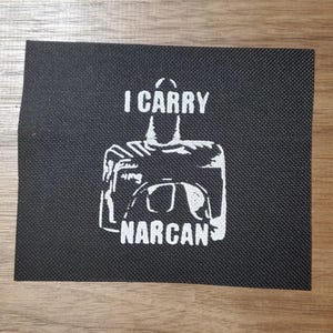 May include: Black rectangular patch with white text and graphic. The text reads "I CARRY NARCAN". The graphic depicts a person with a medical kit. The patch is made of a textured fabric.