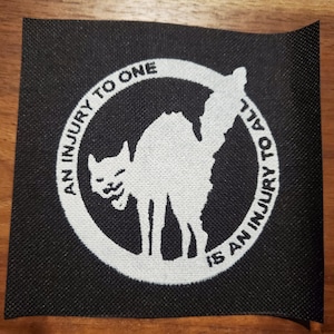 May include: Black and white patch with a circle and a cat silhouette. The text inside the circle reads "An injury to one is an injury to all."
