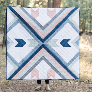 May include: A geometric quilt with a white background and a pattern of blue, gray, and pink triangles and diamonds. The quilt is hanging in a forest setting.