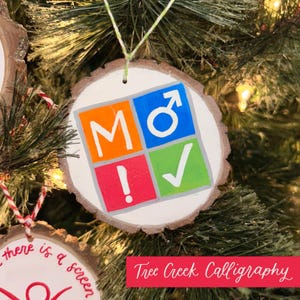 May include: A round, wooden Christmas ornament with a white painted surface. The ornament features the letters "MO" in orange, a male symbol in blue, an exclamation point in red, and a checkmark in green. The ornament is hanging on a Christmas tree.