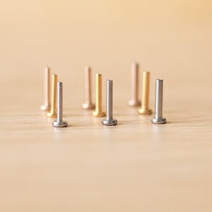 May include: A selection of small stud earrings with flat tops, in gold and silver tones. The earrings are displayed on a light wooden surface, highlighting their minimalist design. The studs are suitable for everyday wear and are measured in millimeters.