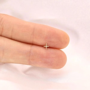 以下が含まれることがあります： 小さな金色の星型のスタッドピアス。ピアスはシンプルな四角い星のデザインです。ピアスは薄いピンク色の背景に表示されています。