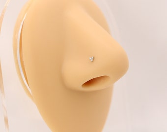 May include: A nose stud featuring three small, clear gemstones arranged in a triangular cluster. The jewelry is set on a silver-colored post and is displayed on a mannequin nose.