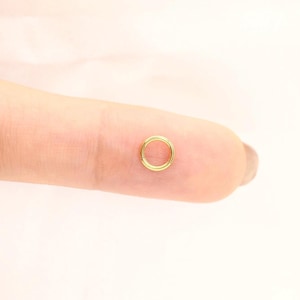 May include: A small, circular gold-toned hoop ring with a smooth, polished finish. The ring features a seamless design and is presented resting on a finger to demonstrate its scale and circular shape, showcasing a simple, continuous metal band.
