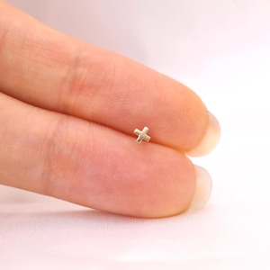 May include: A tiny gold-colored stud earring shaped like a plus sign or cross. The small metal piece features a smooth, polished surface and a simple geometric design, presented as a delicate accessory for ear piercings.