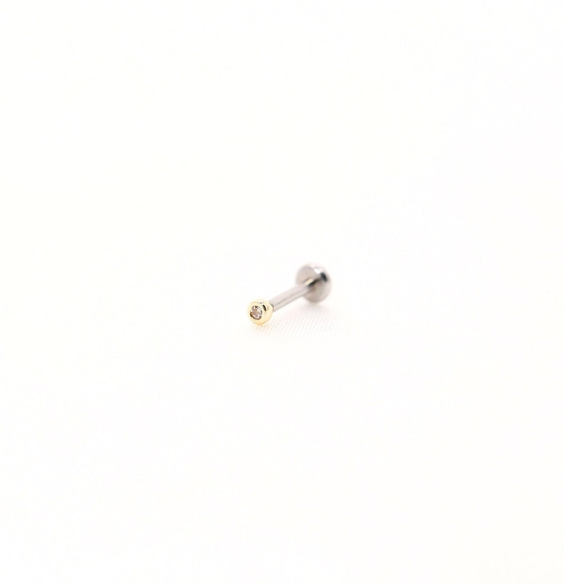 May include: A single, silver-colored labret stud with a small, round, gold-colored bezel setting a tiny, clear gemstone. The stud has a flat back and a ball end. The jewelry is set against a white background.