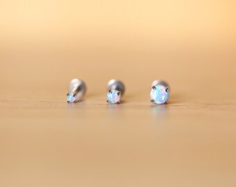 Puede incluir: Tres piezas de joyería corporal de plata con piedras de ópalo. Las piedras son de diferentes tamaños, siendo la más grande la del centro.