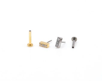 May include: Three gold and silver body jewelry pieces. One is a plain gold post, one is a gold bar with three clear stones, and the other is a silver bar with three clear stones.