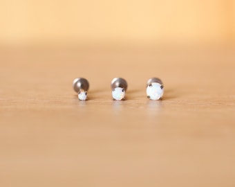 Puede incluir: Tres piezas de joyería corporal de plata con piedras de ópalo blanco. Las piedras son de diferentes tamaños, la más grande a la derecha y la más pequeña a la izquierda.