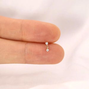 Puede incluir: Un pequeño pendiente dorado con dos gemas redondas y transparentes. Las gemas están conectadas por una delicada cadena, creando un efecto colgante. El pendiente se sostiene entre dos dedos sobre un fondo blanco suave.